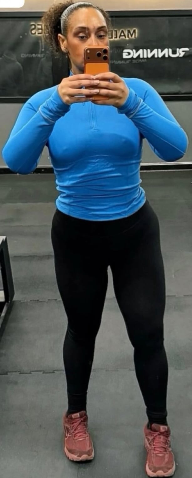Woman in blue long-sleeve top and black leggings taking a mirror selfie at a gym