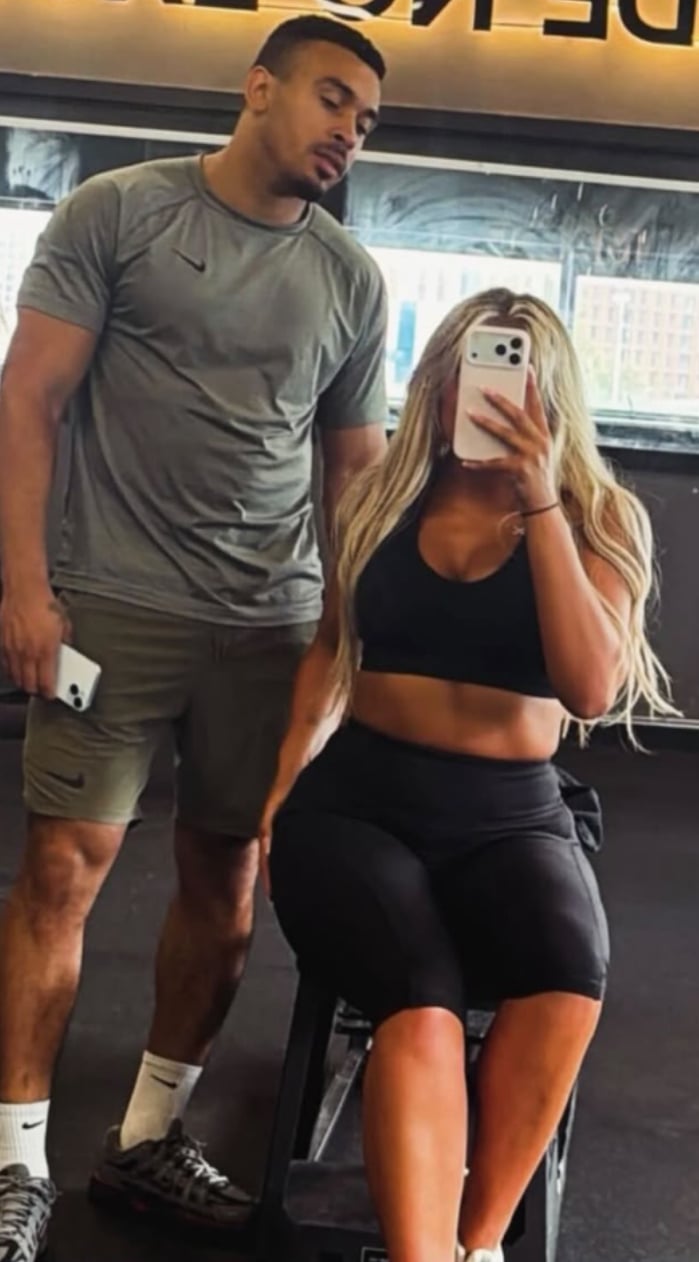 Two people taking a mirror selfie in a gym, man in gray shirt and woman in black sports bra and shorts