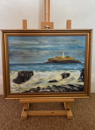 Oil painting on wooden easel depicting a coastal landscape with a lighthouse on a rocky island and sandy beach with dark rock formations