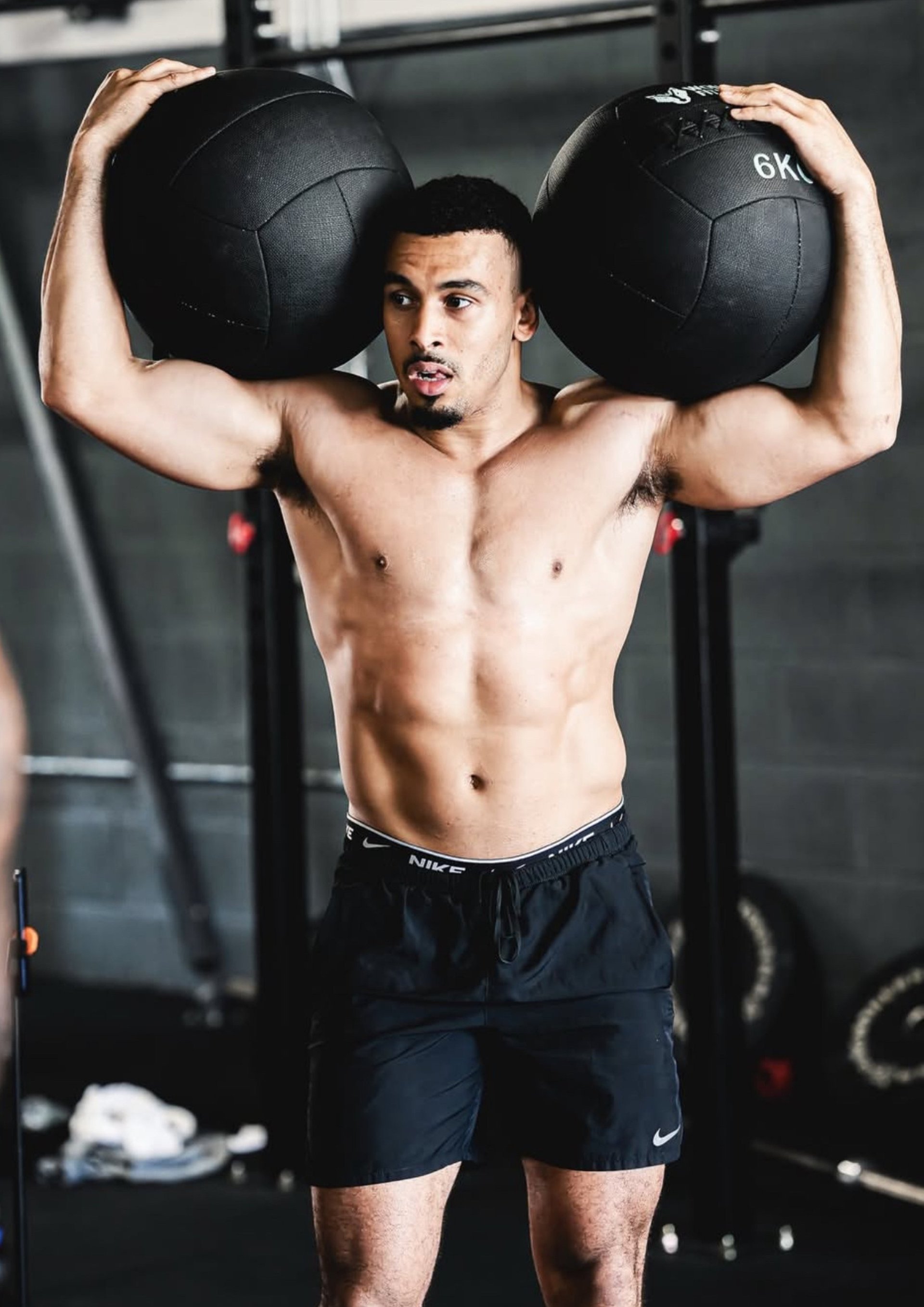 Shirtless athletic man holding two black medicine balls on shoulders in a gym setting
