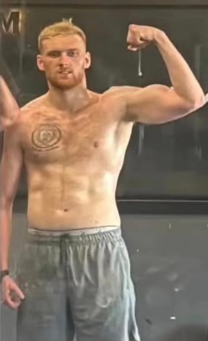 Shirtless man flexing bicep muscle with circular tattoo on chest, wearing gray pants