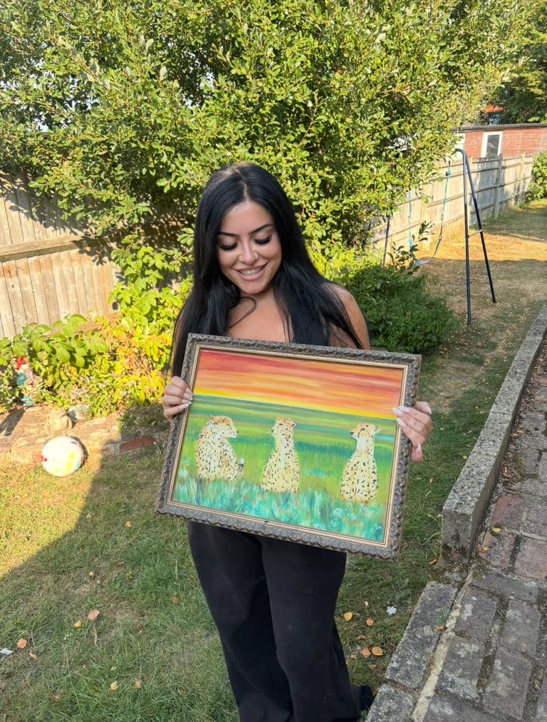 Woman with long dark hair holding a framed artwork featuring dandelions and a colorful sky in a backyard garden setting
