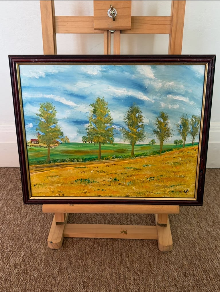 Framed oil painting of a rural landscape with green trees, field, and blue sky displayed on a wooden easel