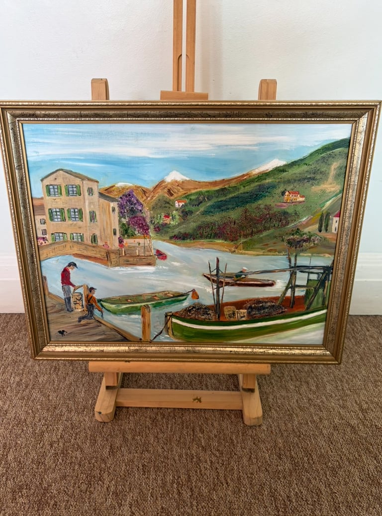 Framed landscape painting on wooden easel depicting Mediterranean coastal village with boats, mountains, and villa architecture