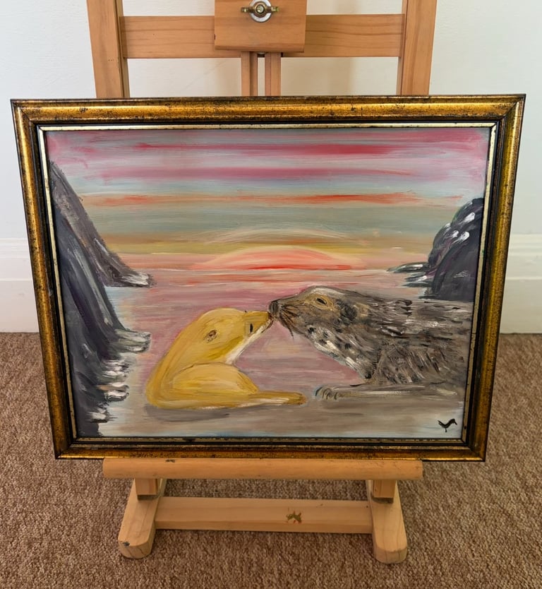 Framed painting on wooden easel depicting two seal-like creatures on a beach at sunset with pink sky and rocky cliffs