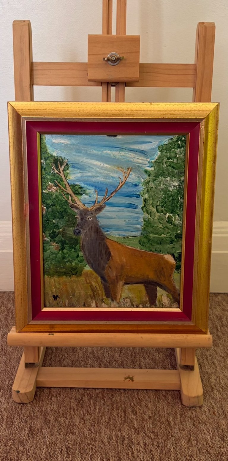 Oil painting of a brown elk with large antlers standing in a forest landscape with blue water and mountains, displayed on a wooden easel with gold and burgundy frame