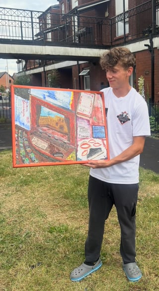 Young man holding a red-framed artwork with colorful collage designs outdoors on grass near brick buildings